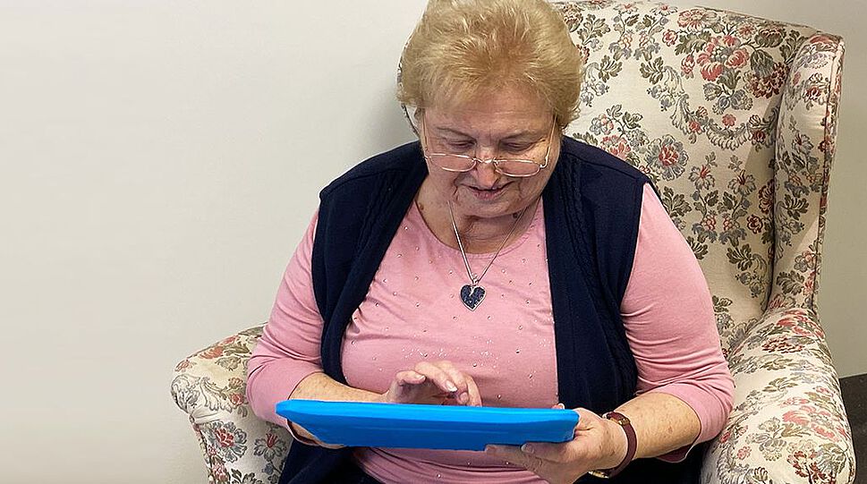Im Bild sitzt eine blonde Seniorin in einem geblümten Ohrensessel und tippt auf einem blauen Tablet. Sie trainiert mit dem Programm von digitAAL Life.
