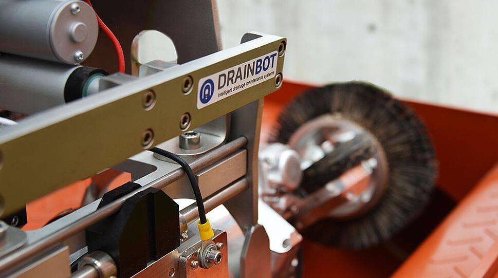 Das Bild zeigt den DrainBot-Roboter in der Ladeposition in einem Drainageschacht. Zu sehen ist ein ca. 70 Zentimeter langer Zylinder, der auf der Vorderseite über eine rotierende Bürste zur Reinigung der Entwässerungsröhren verfügt. Dahinter befindet sich der ca. 25 Zentimeter lange Antriebs- und Sensorikteil des Reinigungsroboters.