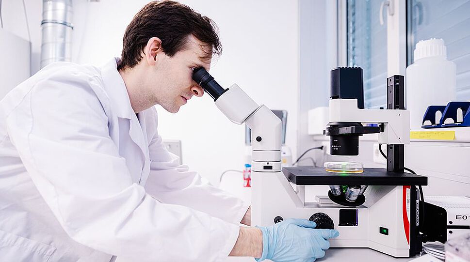 Das Bild zeigt einen Wissenschaftler in weißem Mantel an einem Mikroskop in einem Labor.