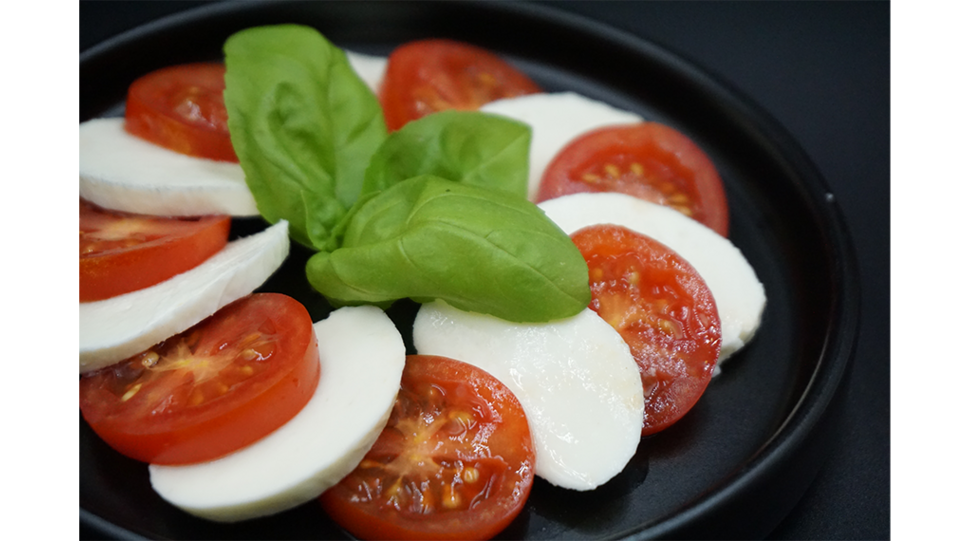 Auf dem Bild ist ein schwarzer Teller mit Tomaten- und Mozzarellascheiben zu sehen, garniert mit drei grünen Basilikumblättern. Der vegane Mozzarella wurde mit Fermify-Proteinen fermentiert. © Fermify