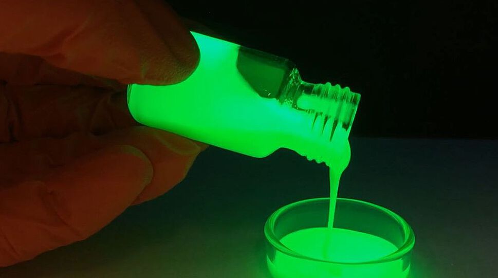 Das Bild zeigt vor dunklem Hintergrund eine leuchtend grüne Flüssigkeit, die mit lichtemittierenden Perowskit-Quantenpunkten (sogenannten PQDs) durchsetzt ist. Sie wird von Hand aus einem Fläschchen in ein ebenso grünes Glas umgefüllt