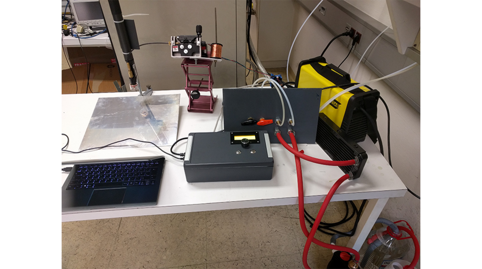 Das Bild zeigt den ersten Versuchsaufbau in seinen Einzelteilen auf einem Schreibtisch. Ein Schweißgerät liefert Strom, ein Panel hält Ventile, Stromanschluss und Wasserkühlung des Brenners, eine eigene Messbox (Mitte unten) zeichnet Daten auf und steuert Motoren, ein krude modifizierter Drahtvorschub ist oben in der Mitte zu sehen. Links oben befindet sich der modifizierte Plasmabrenner, links unten ein Laptop zum Abrufen der Daten. Nicht zu sehen sind Wasserpumpe, Gasflusskontrolle und 5-Achsen-Steuerung. © PluriBot