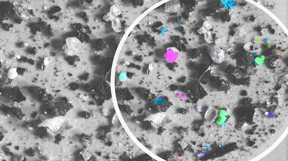 Das Bild zeigt auf einer sonst grauen Fläche mit einzelnen kornartigen Gebilden violett, grün und blau gekennzeichnete Punkte, die zur Verdeutlichung eingekreist sind. Die Software erkennt verlässlich und in wenigen Minuten, welche Partikel aus Mikroplastik bestehen, und markiert sie farblich. Die Farben stehen jeweils für eine spezifische Art von Plastik.